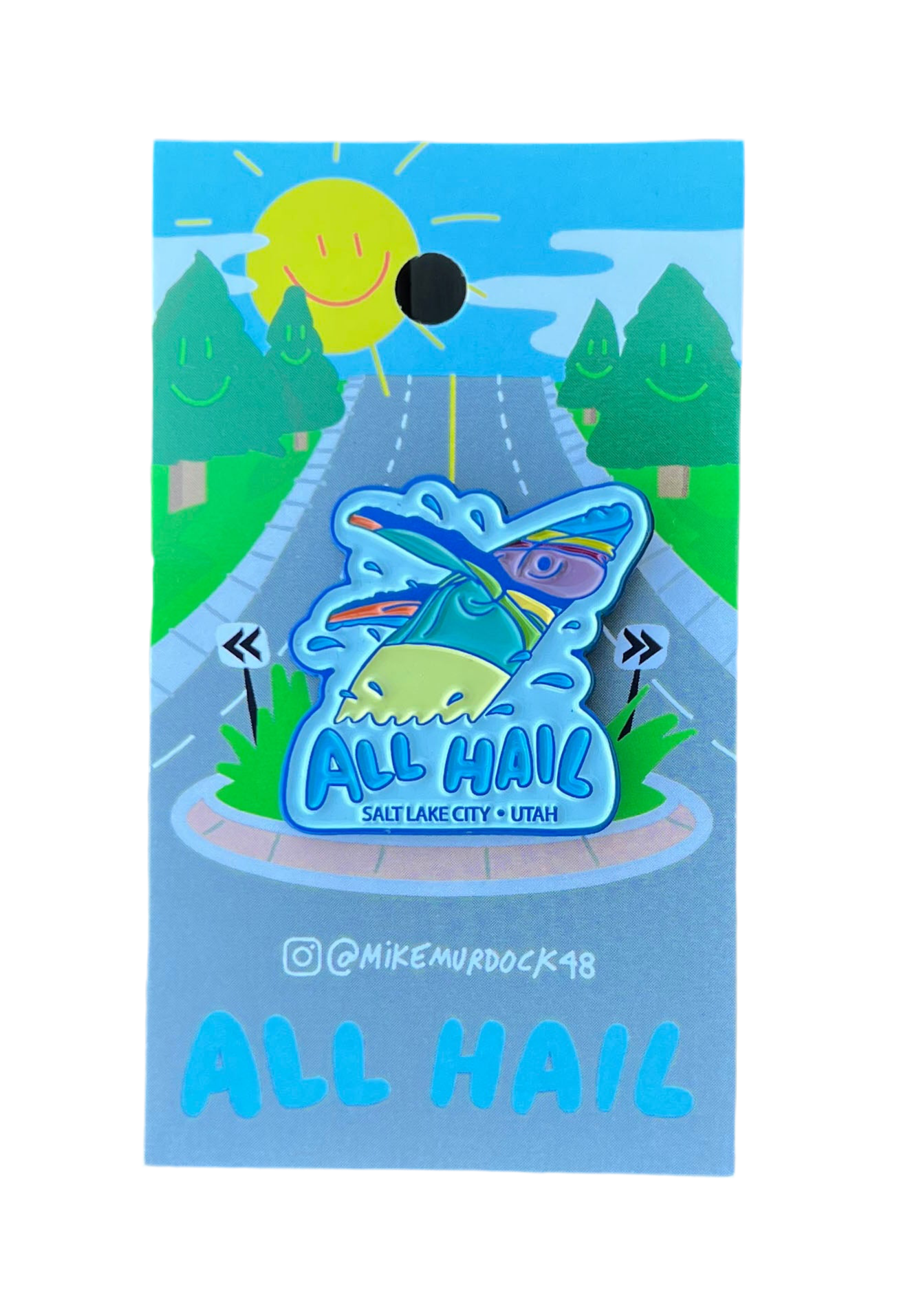 An enamel pin designed with a stylized whale and the text 'All Hail' against a blue background, presented on a card with a sun and road design.