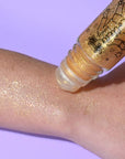 Roll-on bottle of glittery gold cream being applied to a wrist against a purple background