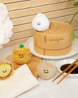 Plush toys resembling dumplings and a steamer basket on a table with a wooden background.