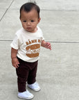 A child modeling a white tee screen printed with brown ink text BANH MI above a smiling banh mi sandwich.