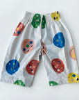 White children's shorts with a colorful abstract print of circles and smiley faces, featuring a green and yellow patterned elastic waistband, laid flat on a vibrant blue background.