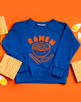 Blue sweatshirt screen printed with orange ink text RAMEN above a bowl of ramen with a smiley face, chopsticks, and soup spoon.