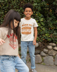A child modeling a white tee screen printed with brown ink text BANH MI above a smiling banh mi sandwich. Another child modeling a tan sweater with Ramen text and smiling bowl of soup in red ink.