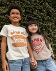 A child modeling a white tee screen printed with brown ink text BANH MI above a smiling banh mi sandwich. Another child modeling a tan sweater with Ramen text and smiling bowl of soup in red ink.