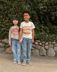 A child modeling a white tee screen printed with brown ink text BANH MI above a smiling banh mi sandwich. Another child modeling a tan sweater with Ramen text and smiling bowl of soup in red ink.