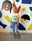 A child modeling a white tee screen printed in green ink text BIBIMBAP above pot of smiling bibimbap, chopsticks, and a spoon.