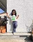 A child modeling a white tee screen printed in green ink text BIBIMBAP above pot of smiling bibimbap, chopsticks, and a spoon.