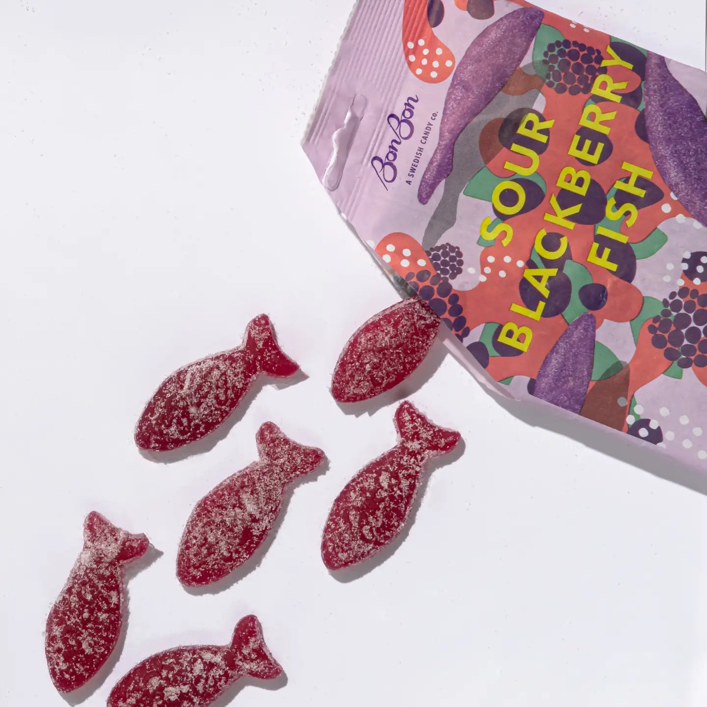 A purple bag of candy with Sour Blackberry Fish written in yellow on the front the bag is open with six purple sour fish gummies spilling out.