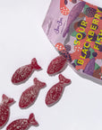 A purple bag of candy with Sour Blackberry Fish written in yellow on the front the bag is open with six purple sour fish gummies spilling out.