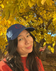 Woman in front of fall tree with yellow leaves wearing a blue hat with a red embroidered smiley face.