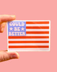 Hand holding a sticker with an American flag design and text 'Could be better' on a pink background
