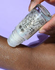 Hand holding a bottle of glittery product applied to skin against a purple background