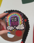 A video of hand holding a vinyl sticker with a sparkly rainbow in the clouds with a smiley face with the words It's Okay To Be Proud.