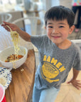 A child modeling a grey tee screen printed with yellow ink text RAMEN above a smiling bowl of ramen with chopsticks, and soup spoon.