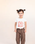 A child modeling a white tee screen printed with smiling mistletoe, snowflakes, mushrooms, leaves, and stars in red.