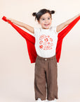 A child modeling a white tee screen printed with smiling mistletoe, snowflakes, mushrooms, leaves, and stars in red.