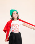 A child modeling a white tee screen printed with smiling mistletoe, snowflakes, mushrooms, leaves, and stars in red.