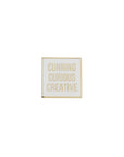 A gold-plated square pin with the text 'Cunning Curious Creative' on it, displayed against a white background.
