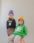 A child modeling a black sweatshirt with screen printed white ink text BOBA above a smiling glass of boba. A second child modeling green sweater with white screen printed Banh Mi text with smiling sandwich.