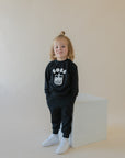 A child modeling a black sweatshirt with screen printed white ink text BOBA above a smiling glass of boba.