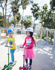 Two children with helmets on riding one red and one green scooter.