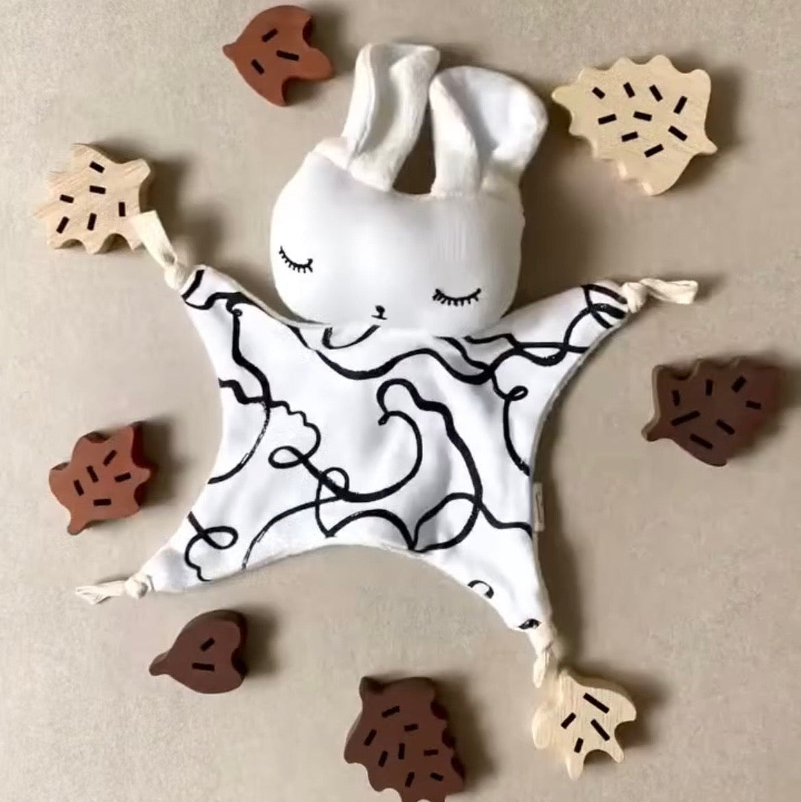 Baby comfort blanket with bunny face surrounded by wooden leaves on a beige background