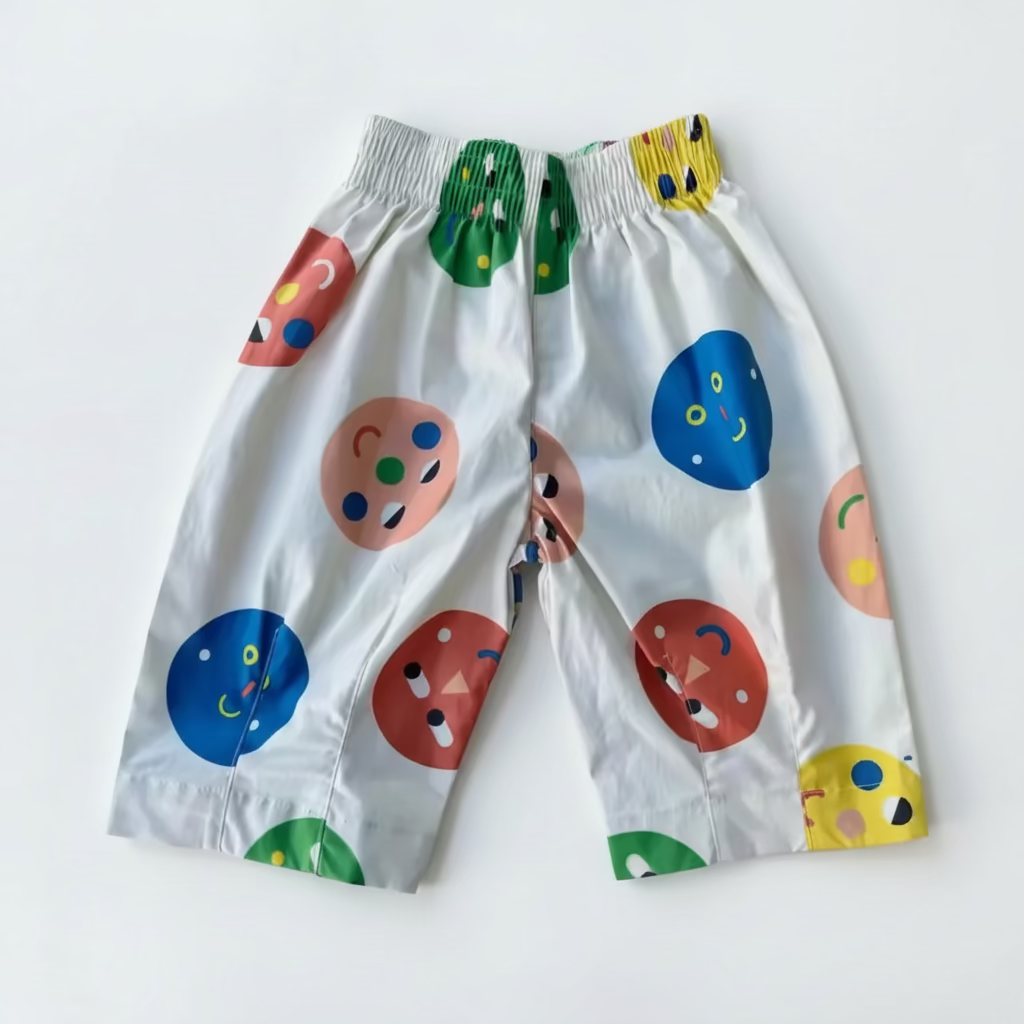 White children's shorts with a colorful abstract print of circles and smiley faces, featuring a green and yellow patterned elastic waistband, laid flat on a vibrant blue background.
