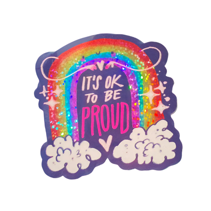 Sparkly  rainbow in the clouds with a smiley face with the words It's Okay To Be Proud.
