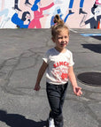 A child modeling a white tee screen printed with red ink, text KIMCHI and cabbage and jar of kimchi, both smiling.
