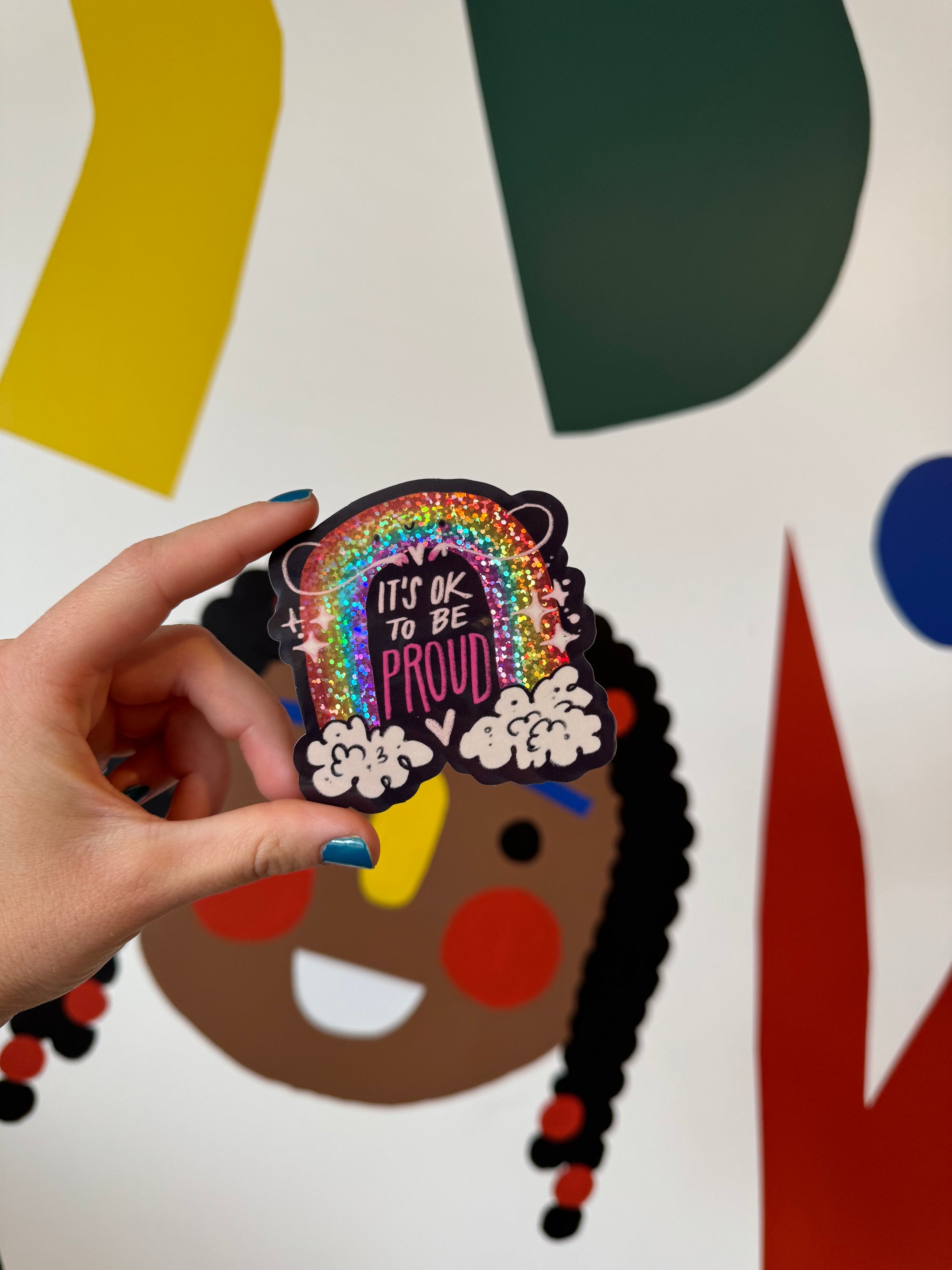 A hand holding a vinyl sticker with a sparkly  rainbow in the clouds with a smiley face with the words It&#39;s Okay To Be Proud.