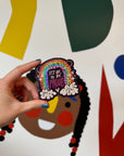 A hand holding a vinyl sticker with a sparkly  rainbow in the clouds with a smiley face with the words It's Okay To Be Proud.