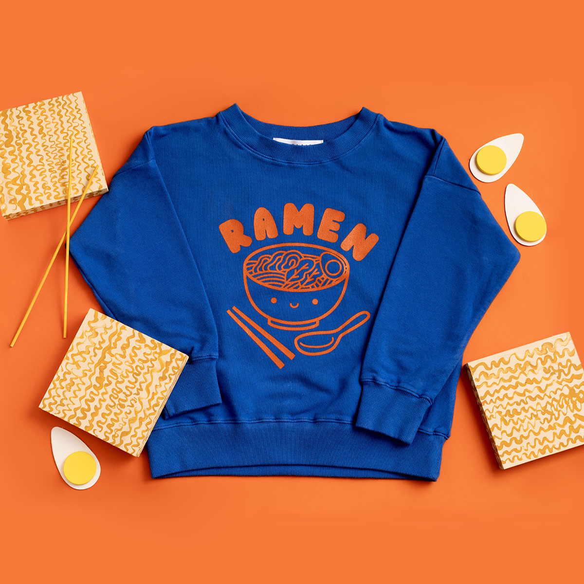 Blue sweatshirt screen printed with orange ink text RAMEN above a bowl of ramen with a smiley face, chopsticks, and soup spoon.