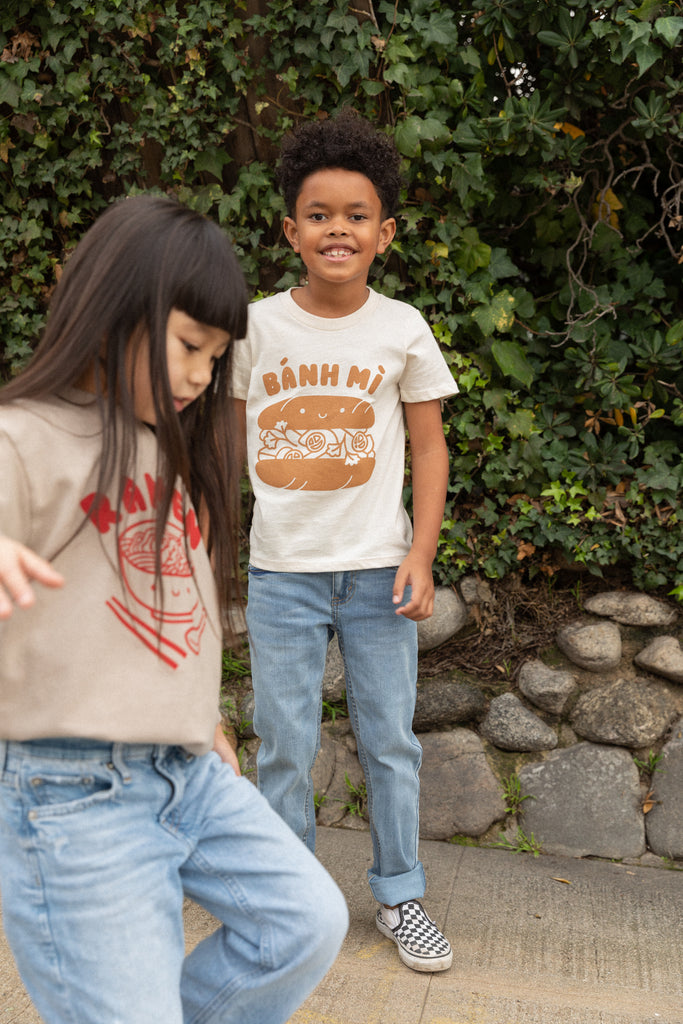 A child modeling a white tee screen printed with brown ink text BANH MI above a smiling banh mi sandwich. Another child modeling a tan sweater with Ramen text and smiling bowl of soup in red ink.