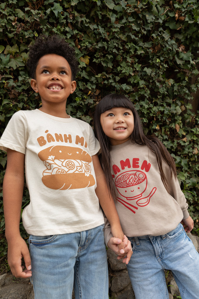 A child modeling a white tee screen printed with brown ink text BANH MI above a smiling banh mi sandwich. Another child modeling a tan sweater with Ramen text and smiling bowl of soup in red ink.