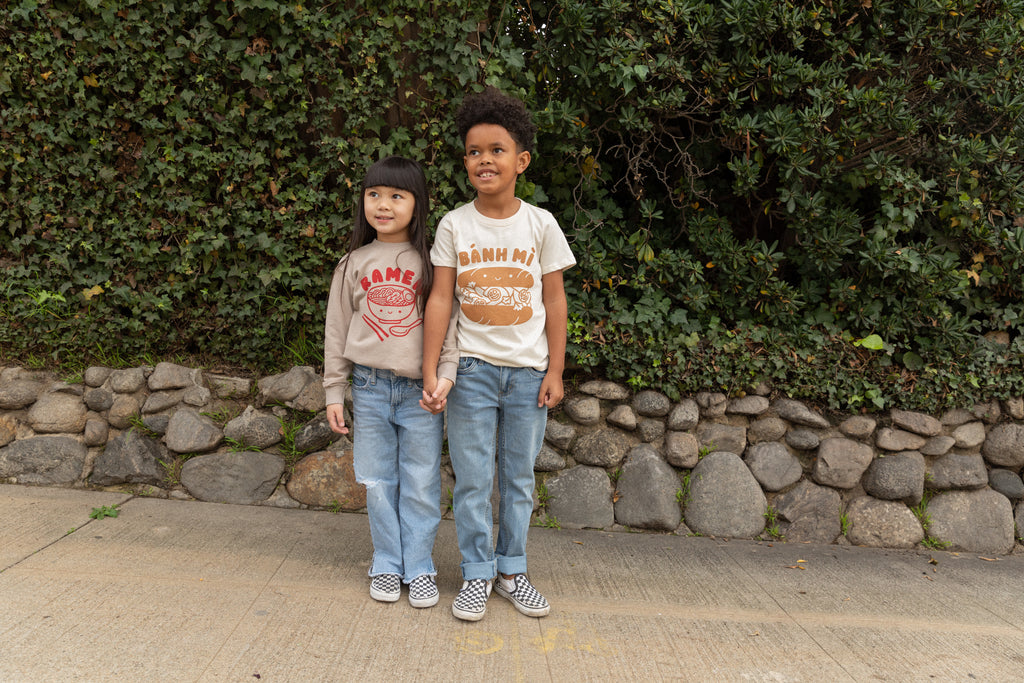 A child modeling a white tee screen printed with brown ink text BANH MI above a smiling banh mi sandwich. Another child modeling a tan sweater with Ramen text and smiling bowl of soup in red ink.