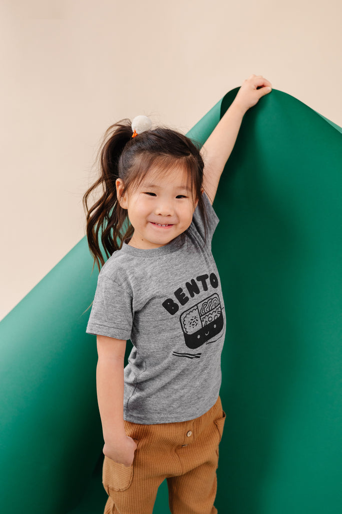 A child modeling a gray tee screen printed with black ink text BENTO above a black smiling bento box lunch with chopsticks.