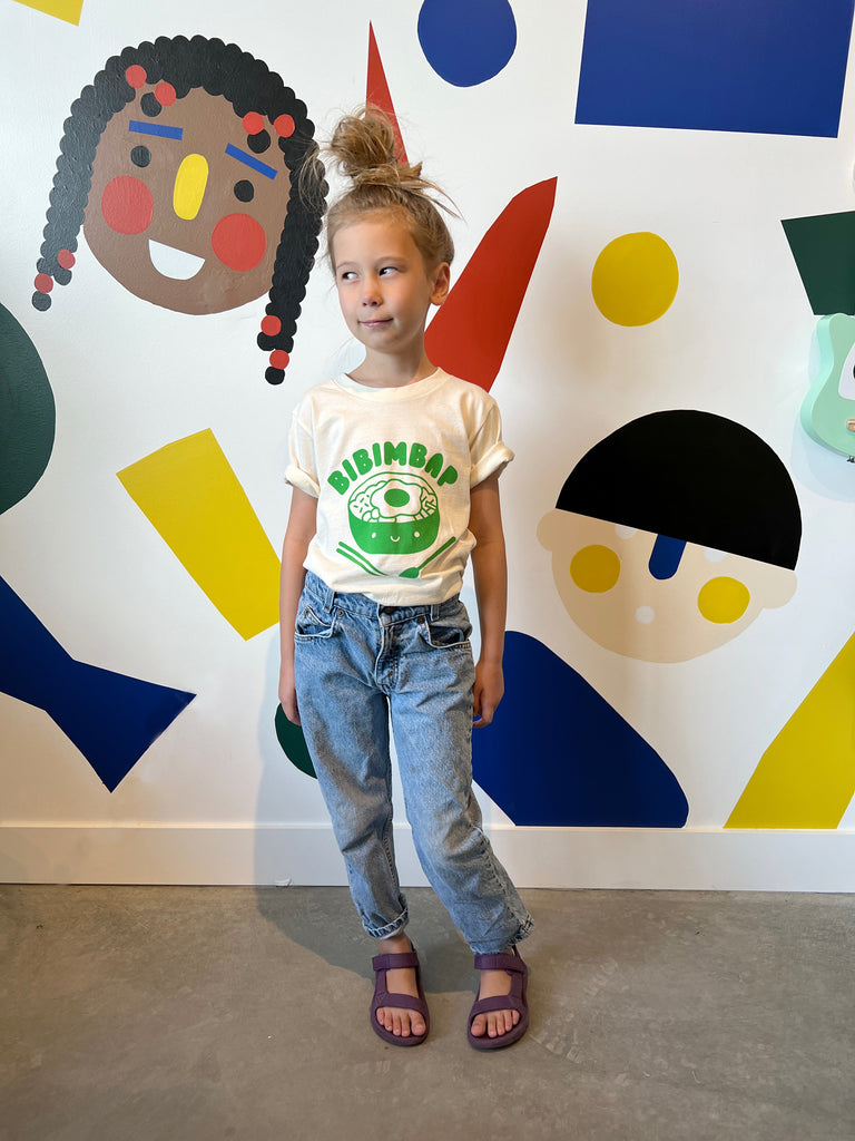 A child modeling a white tee screen printed in green ink text BIBIMBAP above pot of smiling bibimbap, chopsticks, and a spoon.