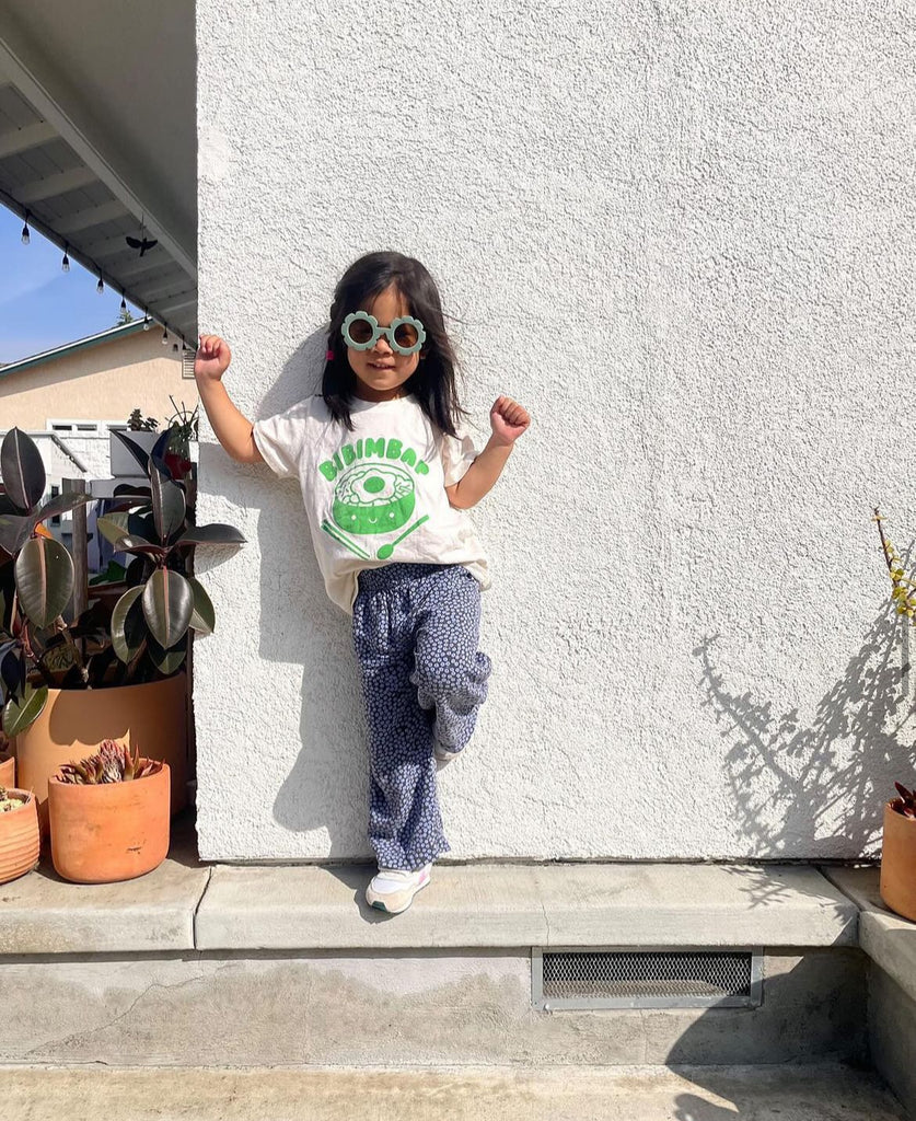 A child modeling a white tee screen printed in green ink text BIBIMBAP above pot of smiling bibimbap, chopsticks, and a spoon.
