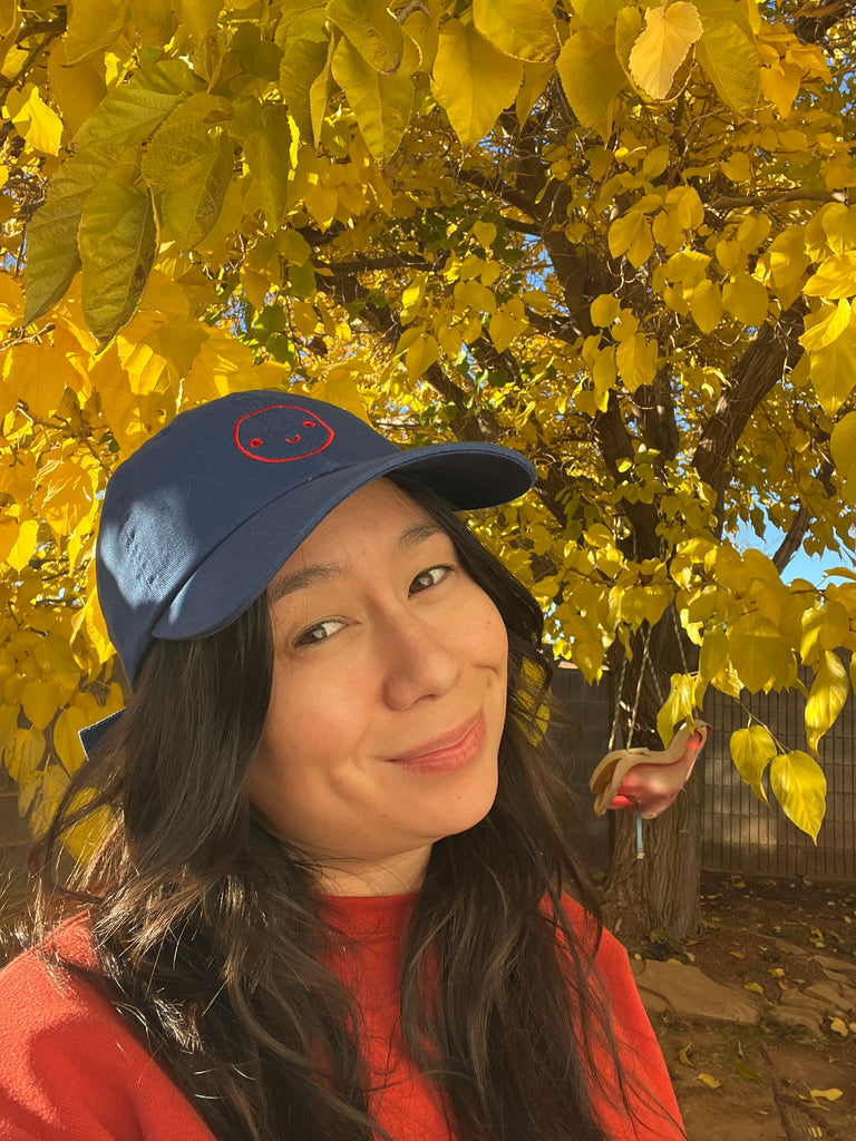 Woman in front of fall tree with yellow leaves wearing a blue hat with a red embroidered smiley face.