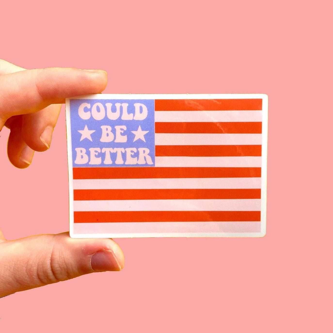 Hand holding a sticker with an American flag design and text 'Could be better' on a pink background