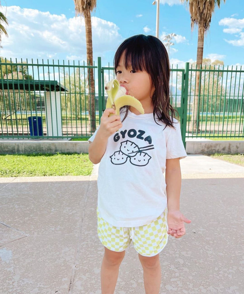 A child modeling a white tee with screen printed black ink text GYOZA above three smiling dumplings.