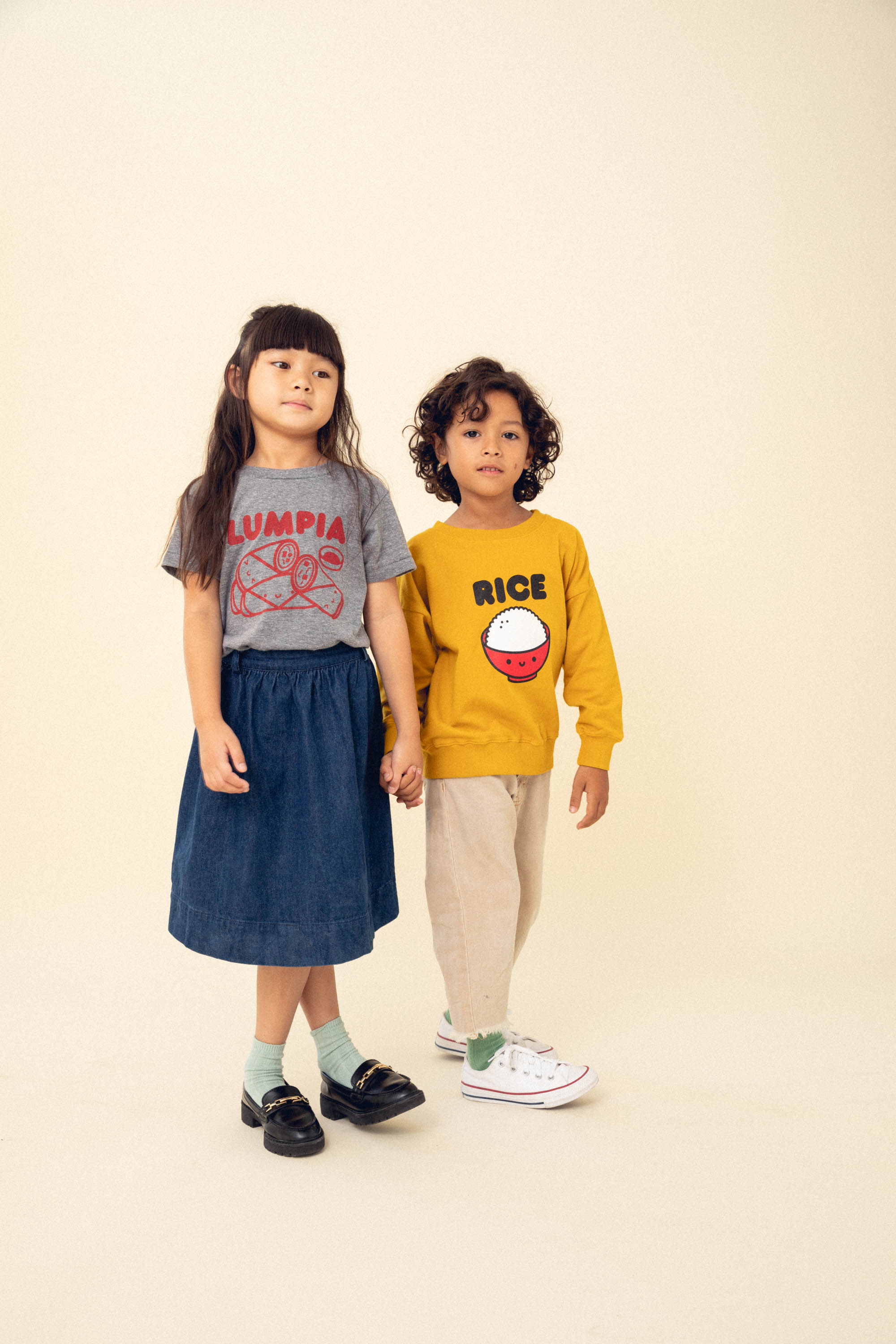 A child modeling a grey tee screen printed with red ink text LUMPIA above three  smiling lumpia and dipping sauce. Another child modeling a yellow sweater with a screen printed red bowl with white rice and the text RICe above in black.