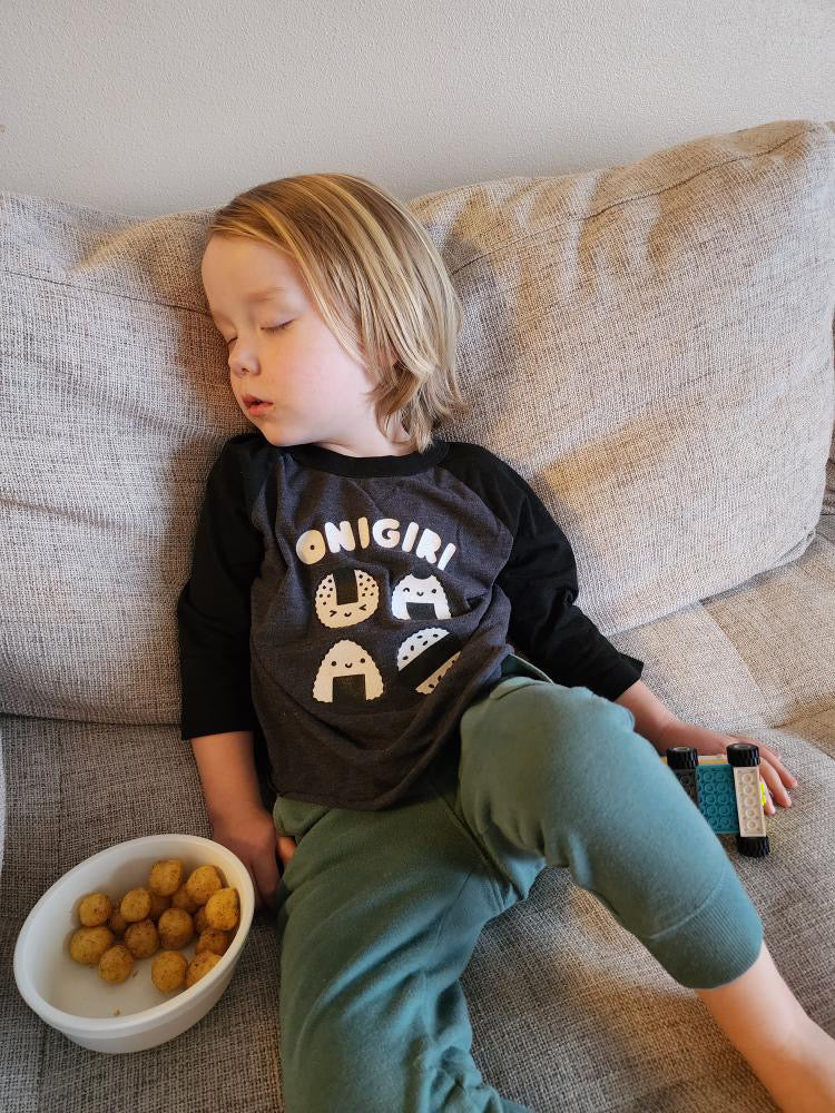 A sleeping child modeling a gray baseball tee with black sleeves printed with white text ONIGIRI above four smiling onigiri.