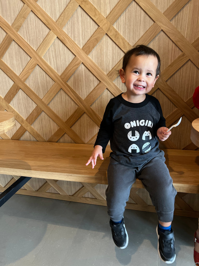 A child modeling a gray baseball tee with black sleeves printed with white text ONIGIRI above four smiling onigiri.
