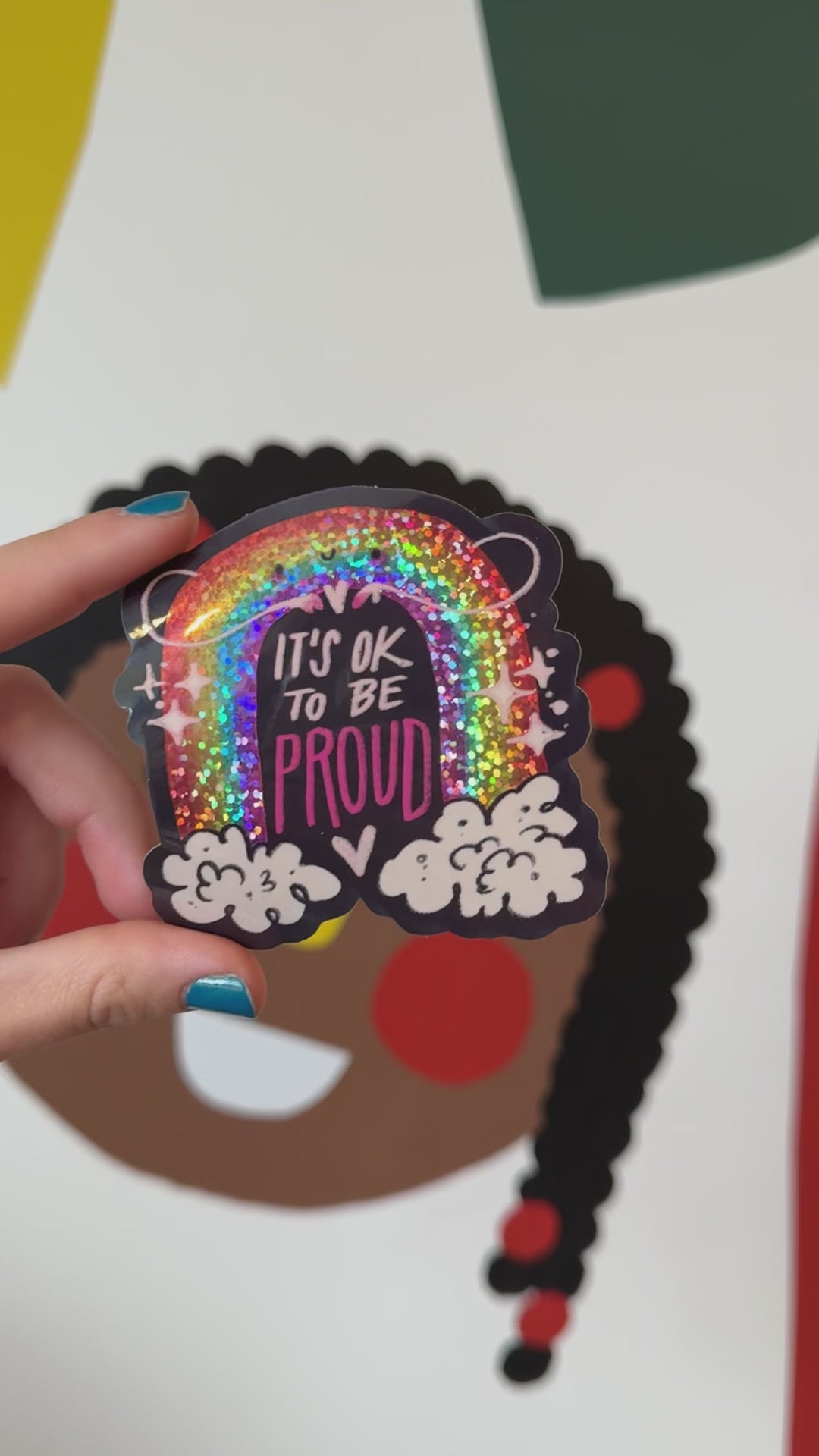 A video of hand holding a vinyl sticker with a sparkly  rainbow in the clouds with a smiley face with the words It's Okay To Be Proud.