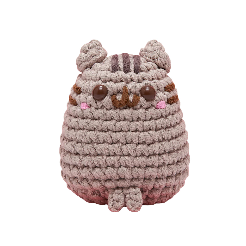 Crocheted plush toy resembling a cat on a pink background, with &#39;Pusheen&#39; branding.