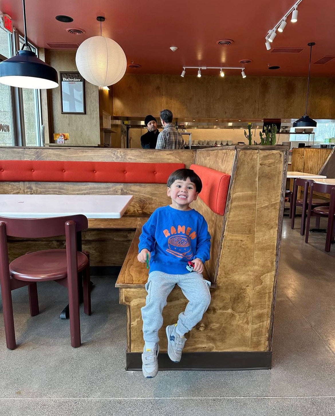 A child modeling a blue sweatshirt screen printed with orange ink text RAMEN above a bowl of ramen with a smiley face, chopsticks, and soup spoon.