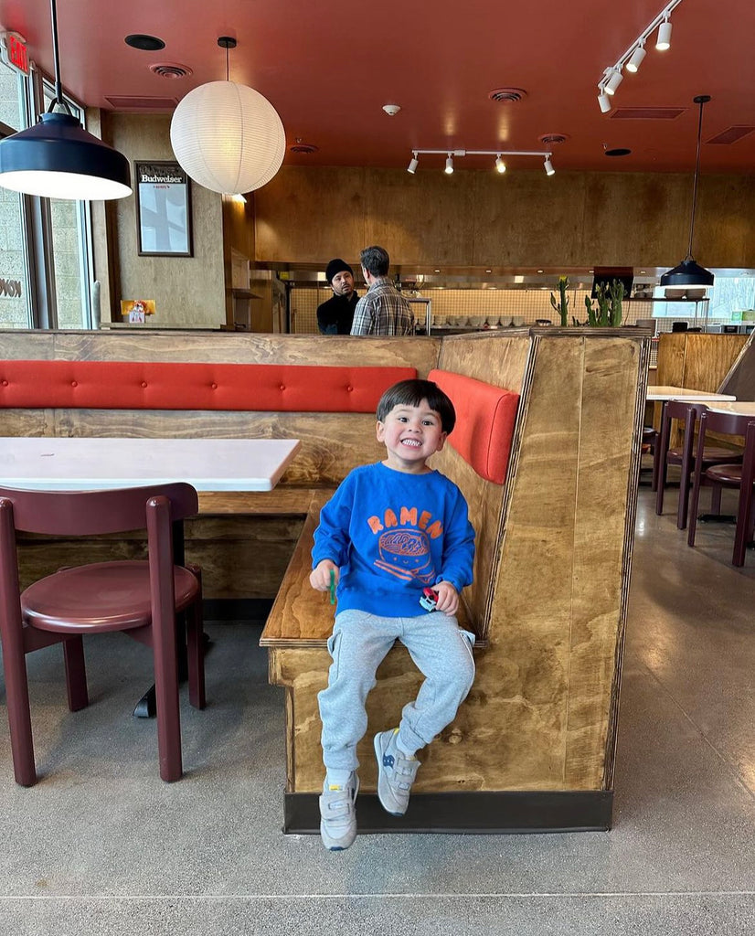 A child modeling a blue sweatshirt screen printed with orange ink text RAMEN above a bowl of ramen with a smiley face, chopsticks, and soup spoon.