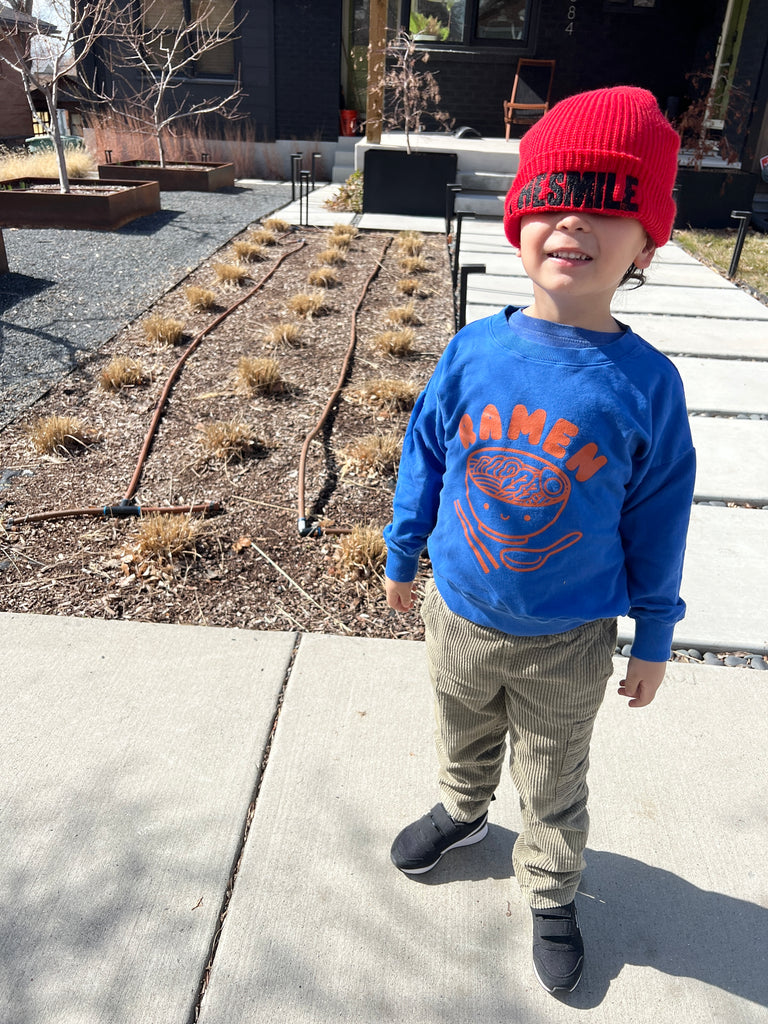 A child modeling a blue sweatshirt screen printed with orange ink text RAMEN above a bowl of ramen with a smiley face, chopsticks, and soup spoon.