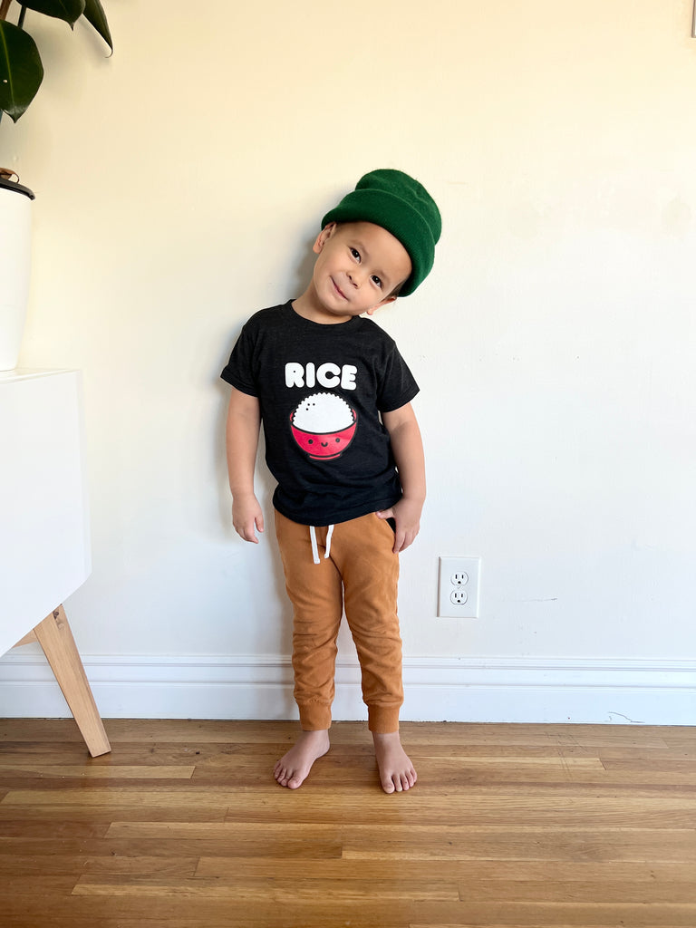 A child modeling a black tee screen printed with white ink text RICE above a red smiling bowl full of white rice.